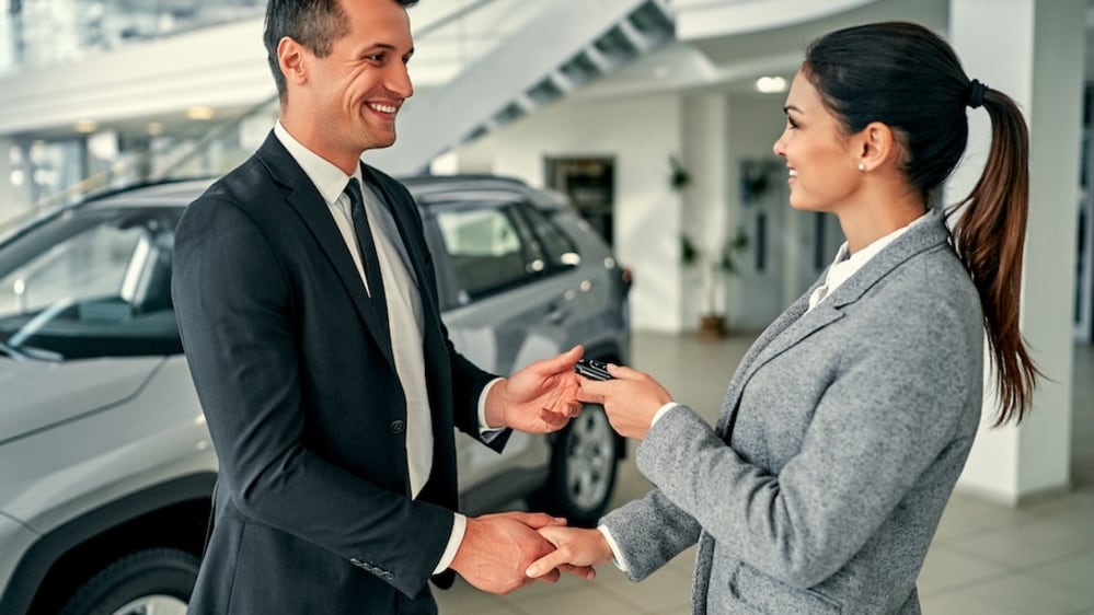 A car dealer handing a customer a car key and shaking hands inside a car dealership