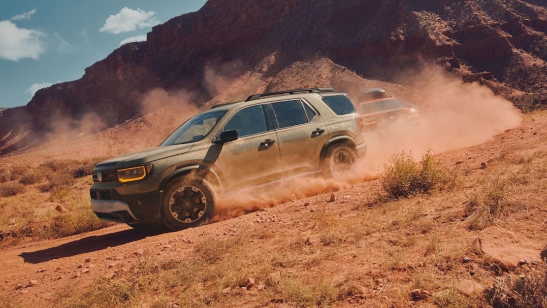 Two Honda SUVs, one olive green and one red, driving down a dusty red-dirt trail