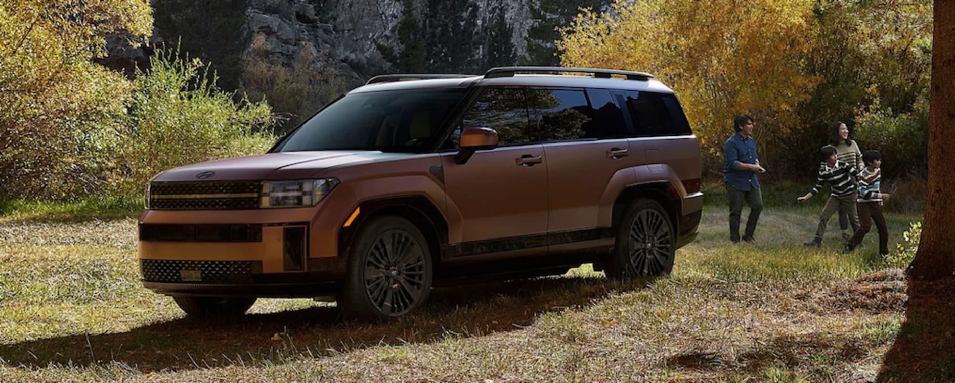 A side view of a copper 2026 Hyundai Santa Fe SUV parked near a mountain, with a family playing a game nearby