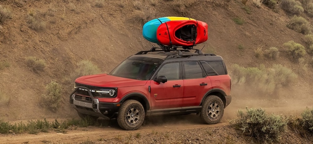 A red 2026 Ford Bronco Sport SUV with boats on its roof rack driving down a dirt road