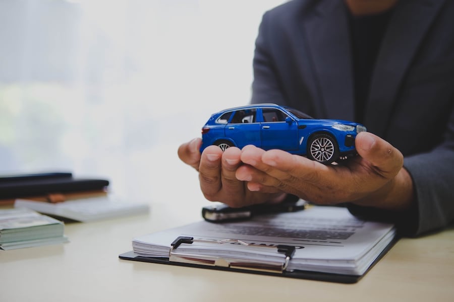A brown-skinned person in a blue suit holding a blue toy car with a clipboard of paperwork and a car key on the desk below their hands