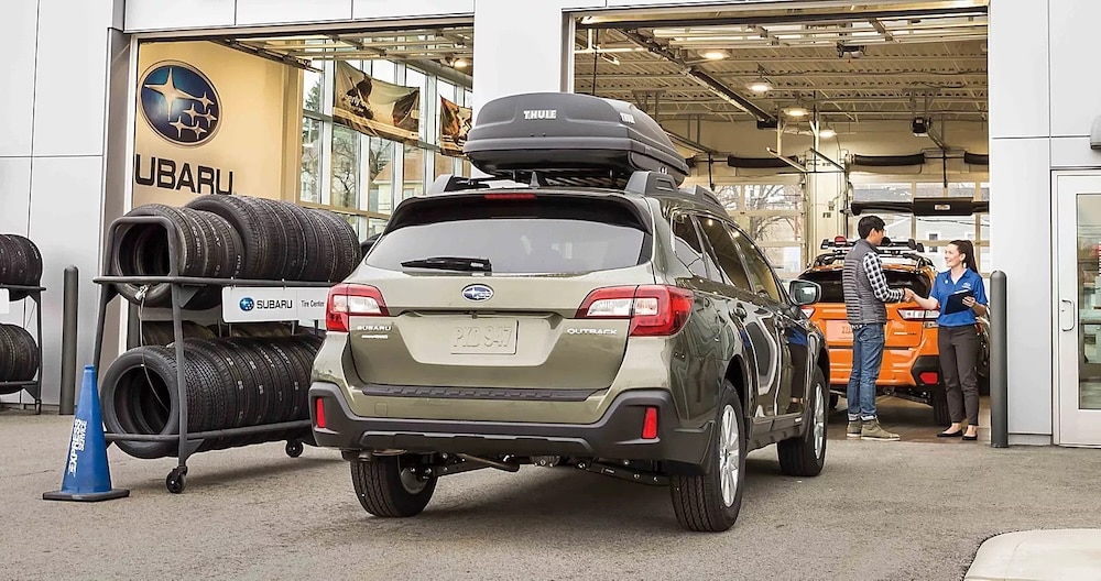 A green Subaru Outback parked in front of a Subaru service center, with a service advisor greeting a customer