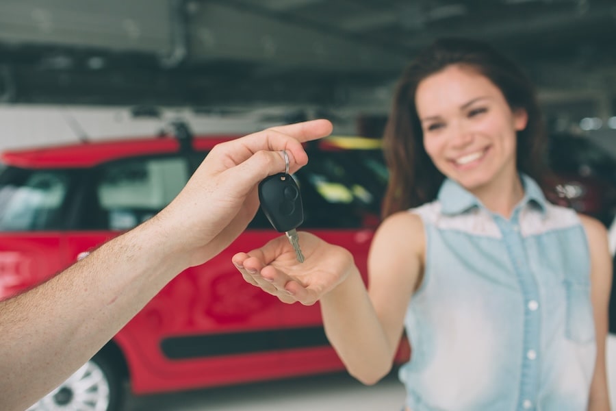 A person handing a smiling woman a car key with a red vehicle in the background