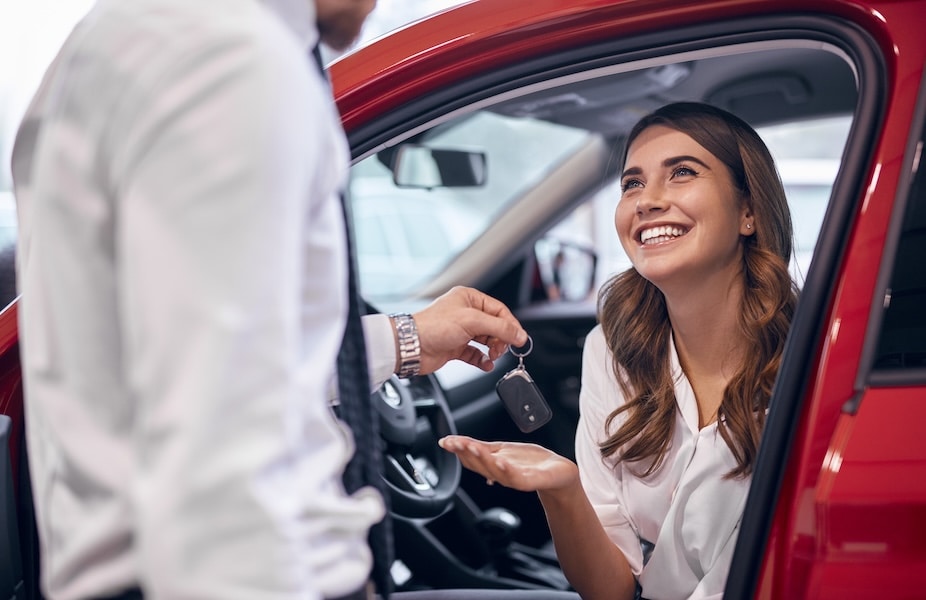 A car salesperson handing a car key to a smiling customer sitting in a red car