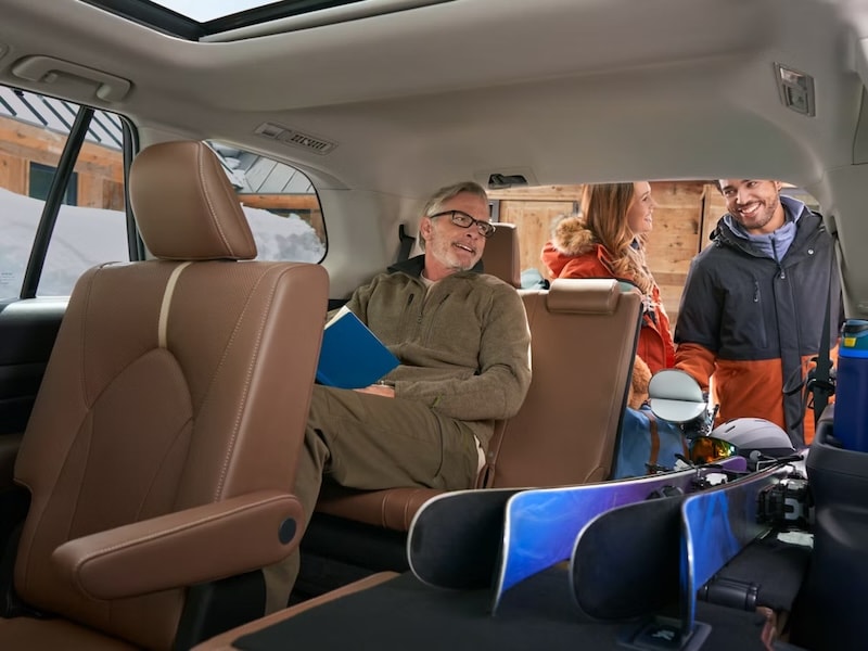 A white man with a beard sitting in the 3rd row of a 2026 Toyota Grand Highlander, while a white woman and another man in snow gear load skis into the rear storage area