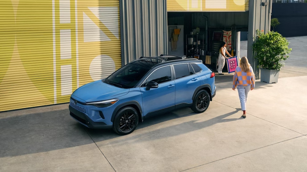 A side view of a bright blue 2026 Toyota Corolla Cross Hybrid SUV parked in front of an art studio