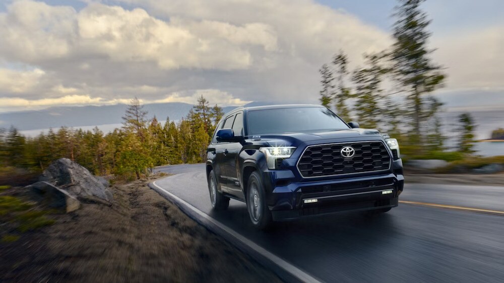 A front view of a dark blue 2026 Toyota Sequoia driving down a winding road, with a lake and mountains in the background