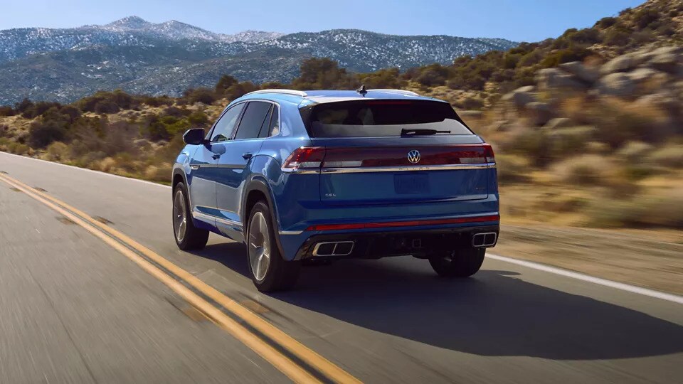 A three-quarters view of the back of a blue 2026 Volkswagen Atlas Cross Sport SUV driving down a mountain desert road