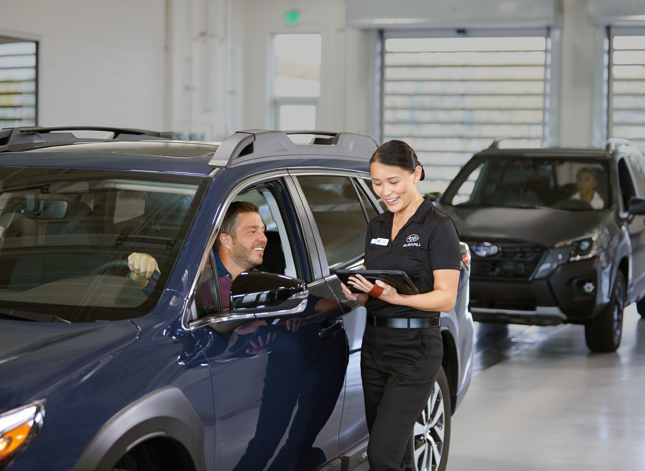 Subaru service advisor reviewing a tablet with a customer seated in a blue Subaru in a service drive
