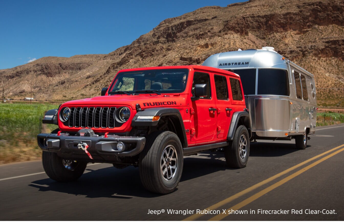 Front three-quarter view of a 2026 Jeep Wrangler Rubicon X in Firecracker Red Clear-Coat towing an Airstream trailer on a highway.
