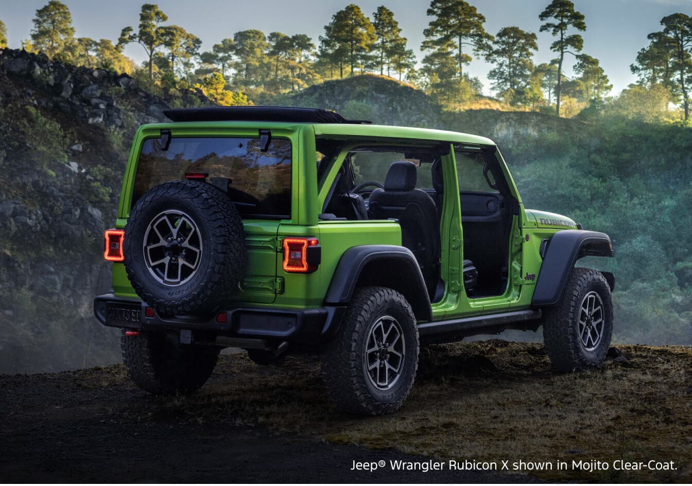 Rear three-quarter view of a 2026 Jeep Wrangler Rubicon X in Mojito Clear-Coat parked on a dirt trail in a dense forest.