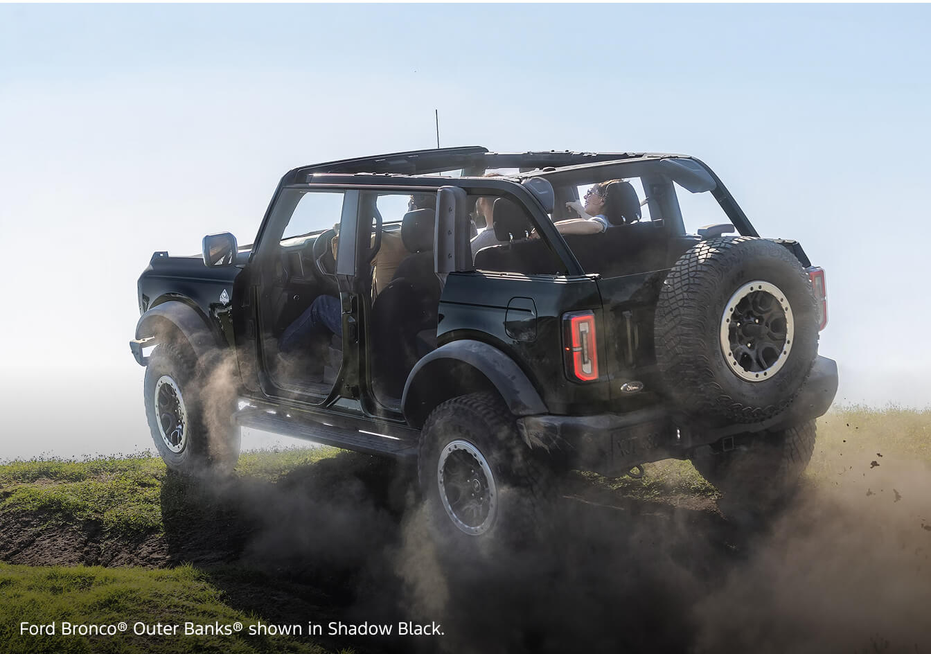 Rear three-quarter view of a 2026 Ford Bronco Outer Banks in Shadow Black kicking up dust while driving open-top over a grassy hill.