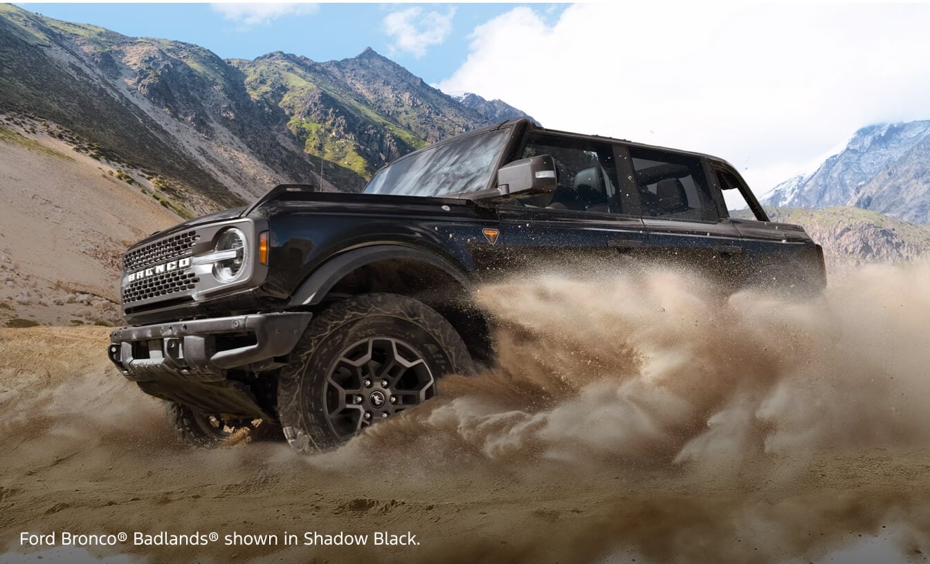 Front three-quarter view of a 2026 Ford Bronco Badlands in Shadow Black carving through sand dunes with mountains behind.
