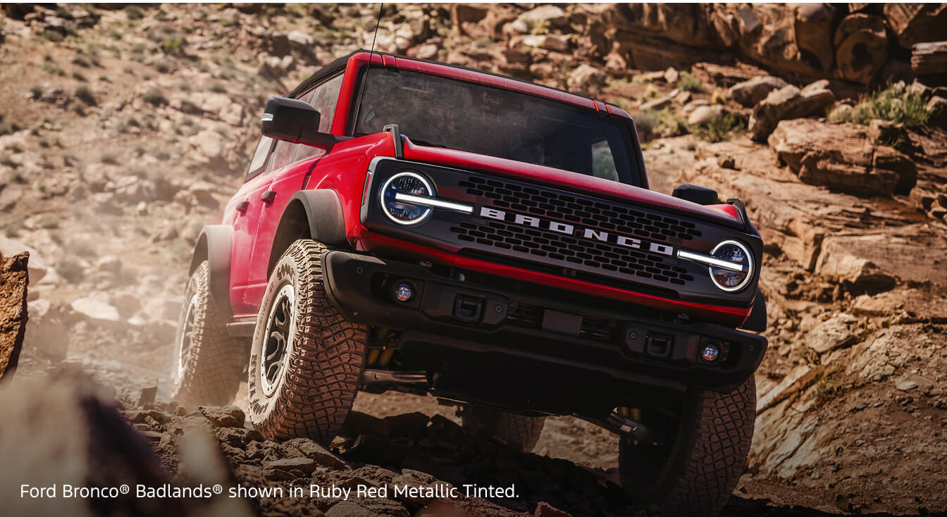 Low-angle view of a 2026 Ford Bronco Badlands in Ruby Red Metallic Tinted Clearcoat traversing rocky terrain in a desert canyon.