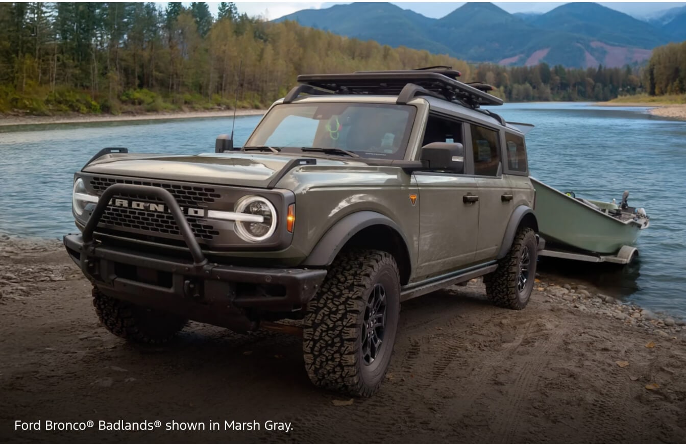 Front three-quarter view of a 2026 Ford Bronco Badlands in Marsh Gray launching a small boat at a river bank with mountains in the background.