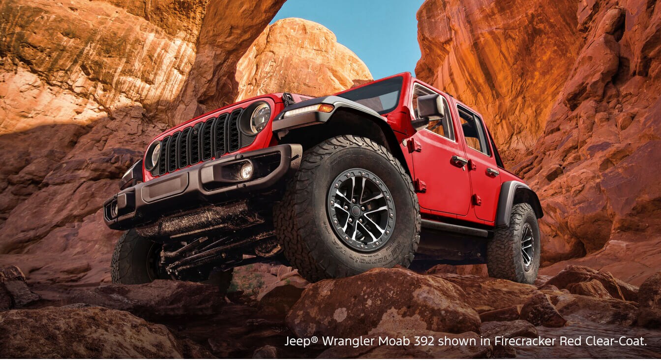 Low-angle view of a 2026 Jeep Wrangler Moab 392 in Firecracker Red Clear-Coat crawling over rocks in a red canyon setting.