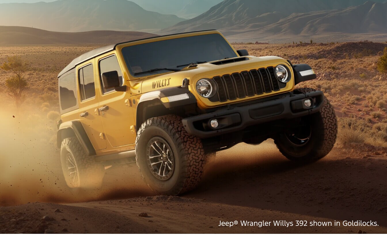 Front three-quarter view of a 2026 Jeep Wrangler Willys 392 in Goldilocks kicking up dust while driving off-road in a desert valley.