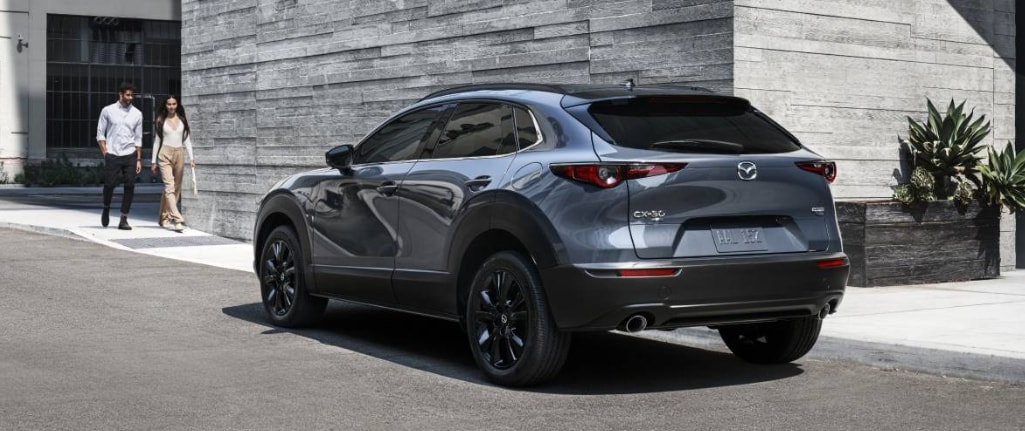 Gray Mazda CX-30 SUV parked by a modern building as a couple walks nearby.