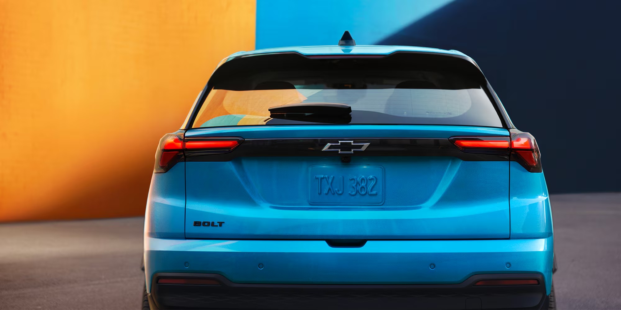 Rear view of a blue 2027 Chevy Bolt in front of an orange and blue wall