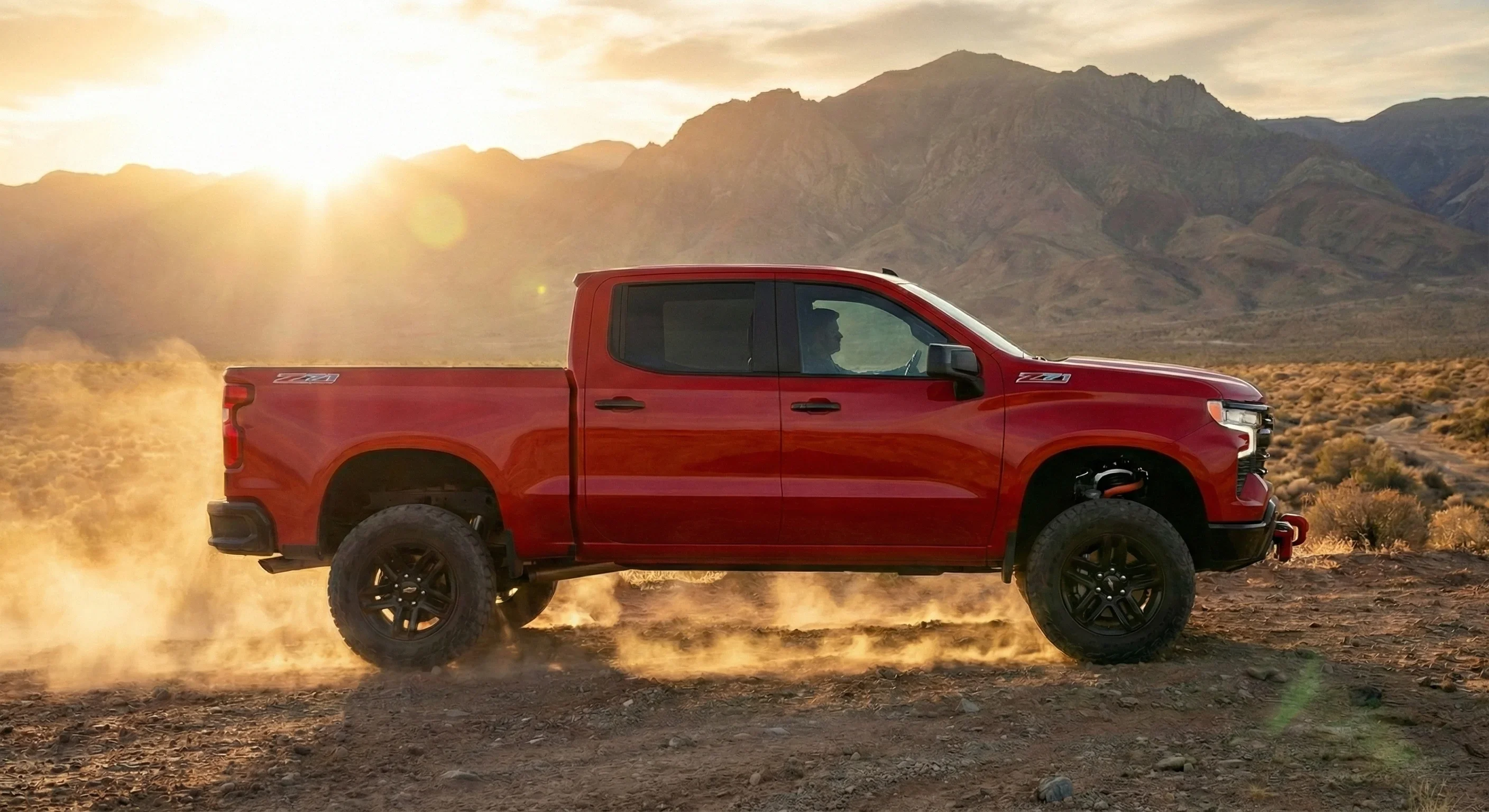 Side view of a red Chevy Silverado Trail Boss with red recovery hooks driving on a dusty desert trail