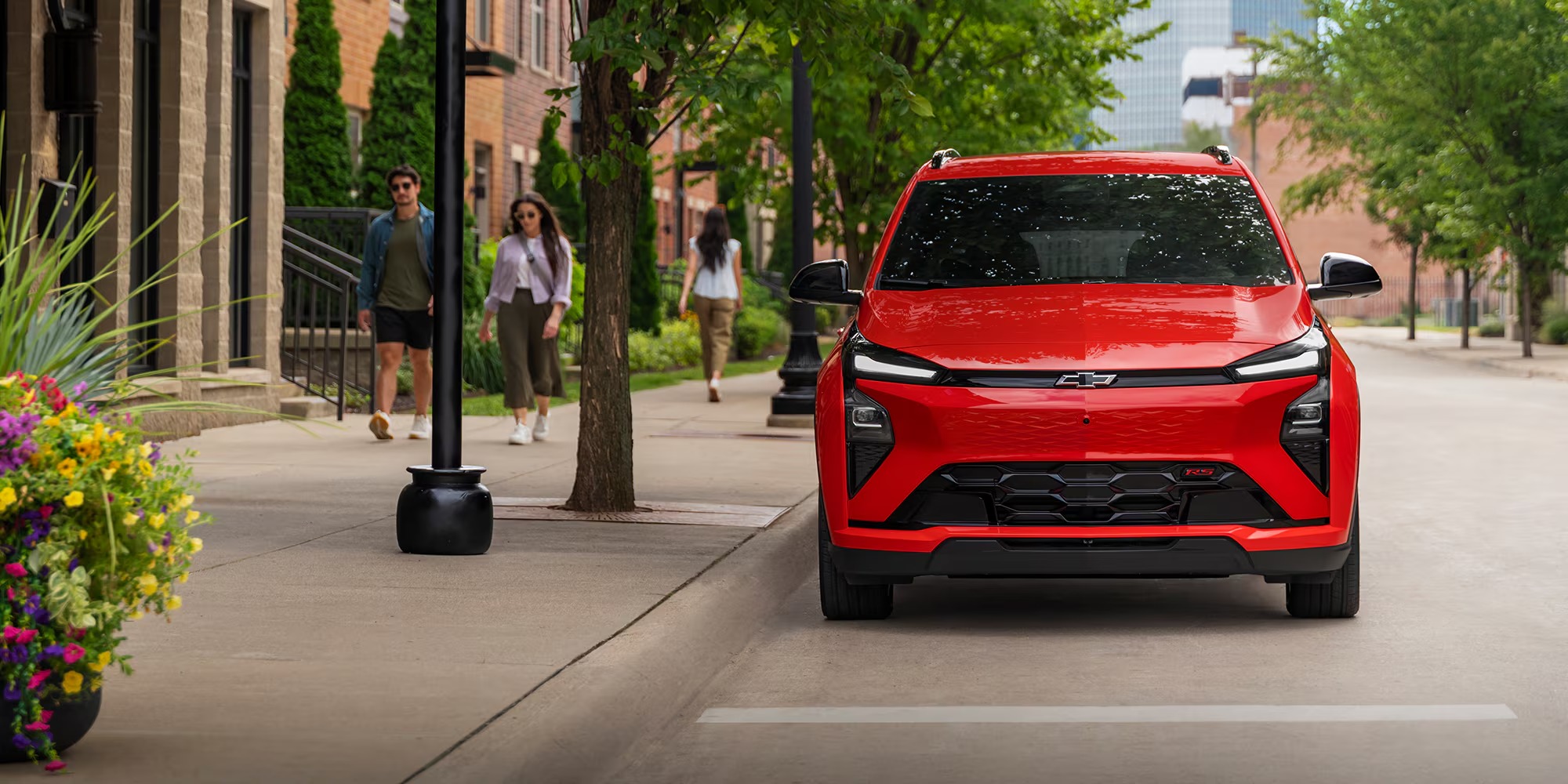 Front view of a red Chevy Bolt with a black grille on a tree-lined downtown street