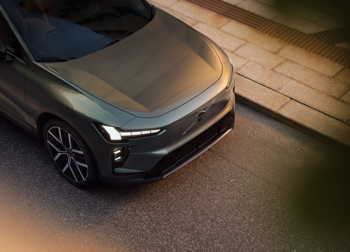 High-angle front view of a green 2026 Volvo EX60 parked on a street next to a sidewalk at sunset.