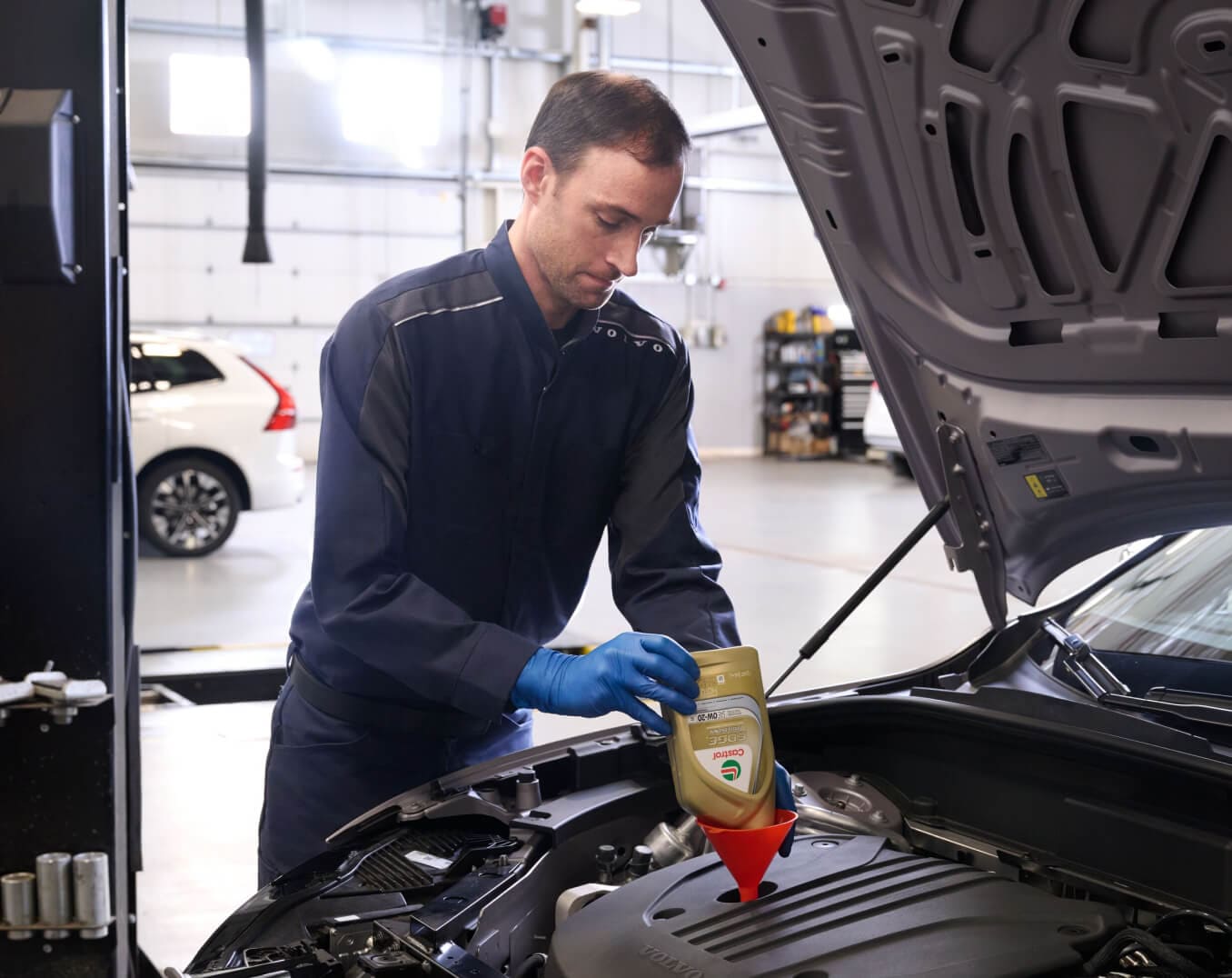 Side view of a technician pouring motor oil into a Volvo car engine in a service bay.