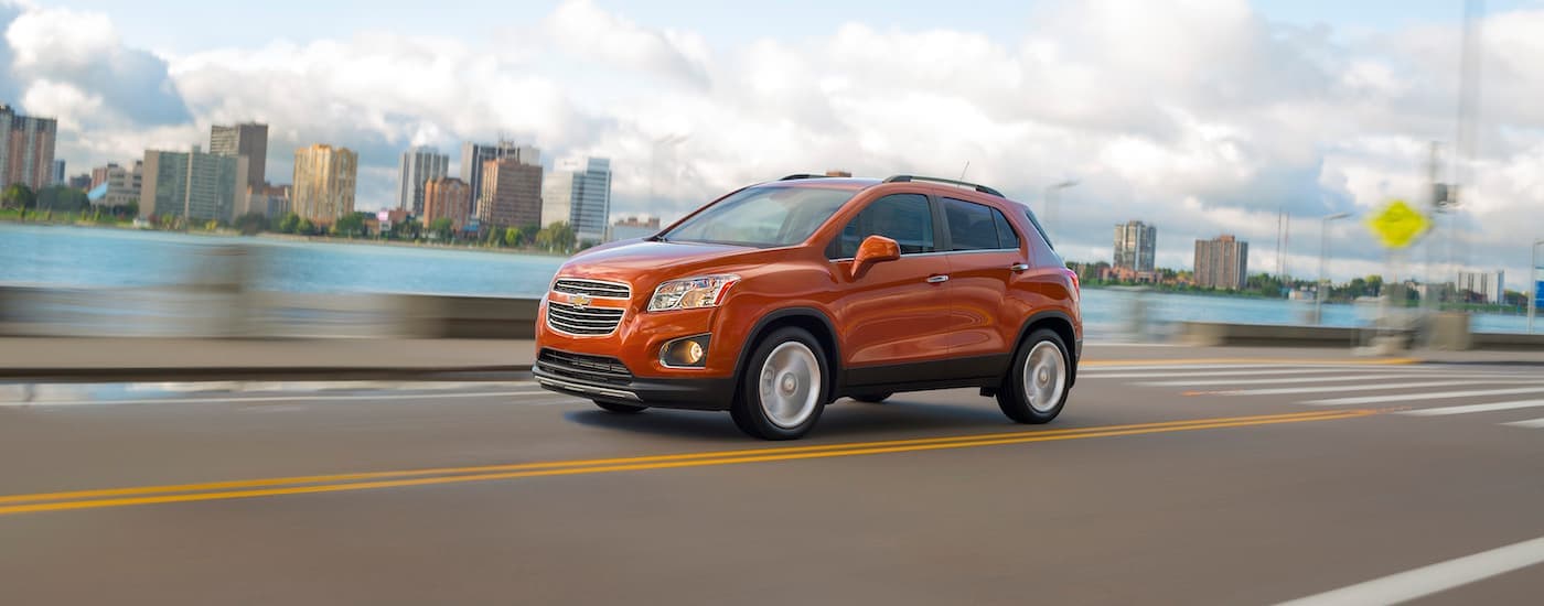 An orange 2015 Chevy Trax for sale is shown driving past city buildings near Columbus, OH.