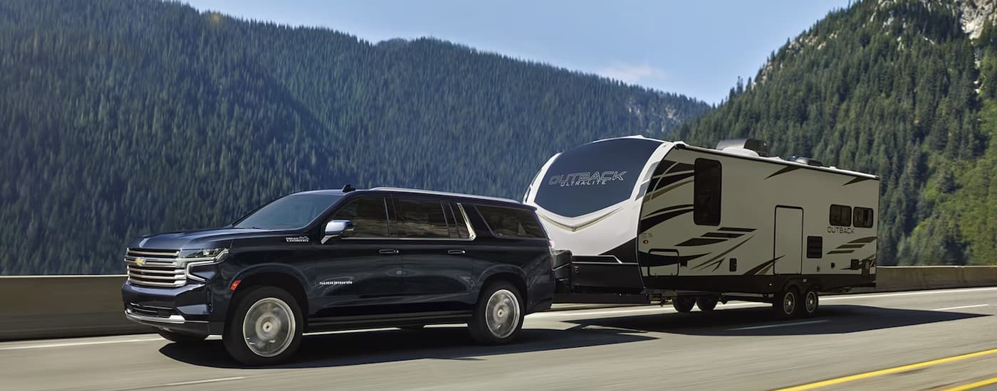 A dark blue 2024 Chevy Suburban High Country is towing a camper trailer on a mountain highway.