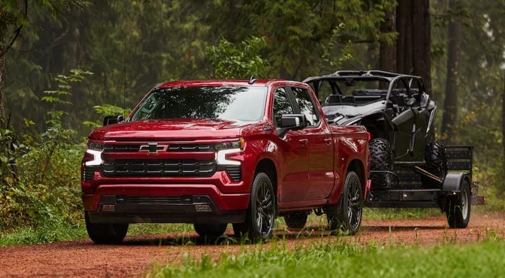 A red 2025 Chevy Silverado 1500 RST is shown towing a trailer off-road.