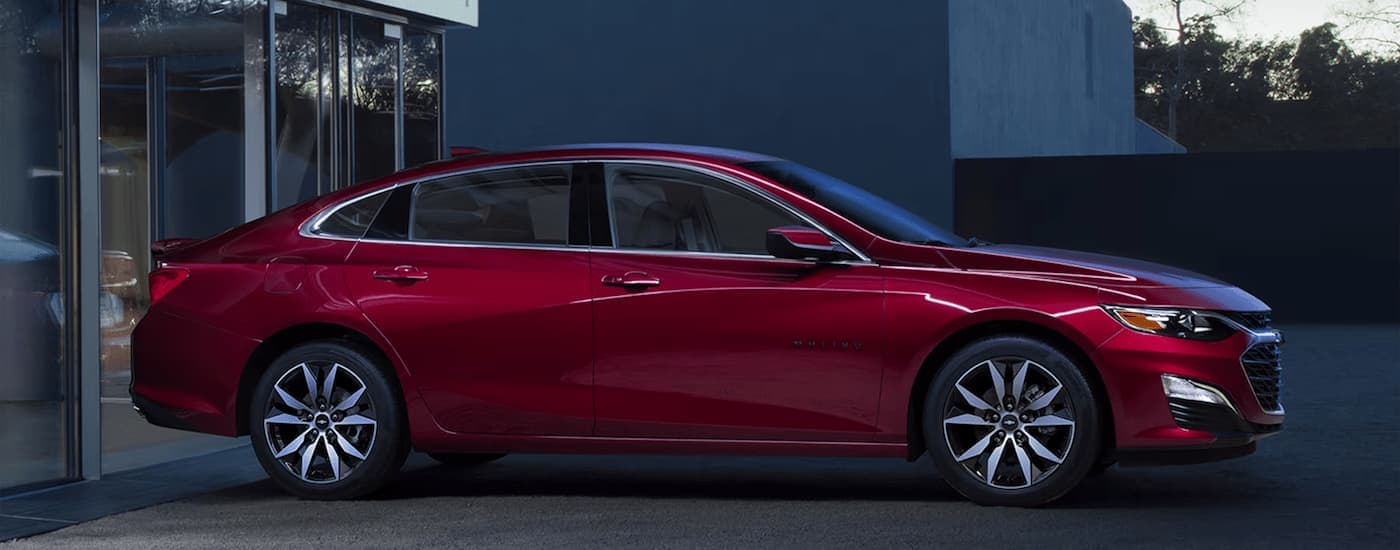 A red 2024 Chevy Malibu Sport is shown from the side parked outside of a Chevy dealer.