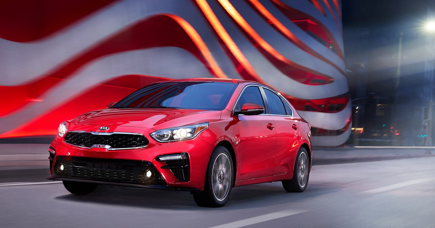 red Kia Forte car driving in front of a curved white and red sculpture