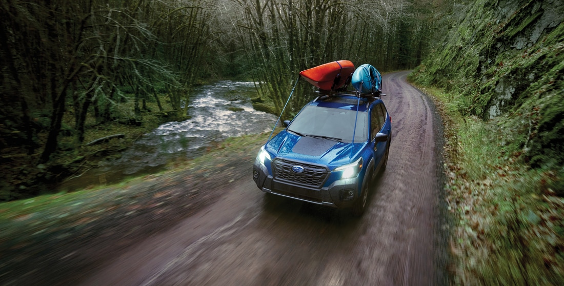 blue Subaru Forester SUV driving down a dirt trail with kayaks on the roof rack