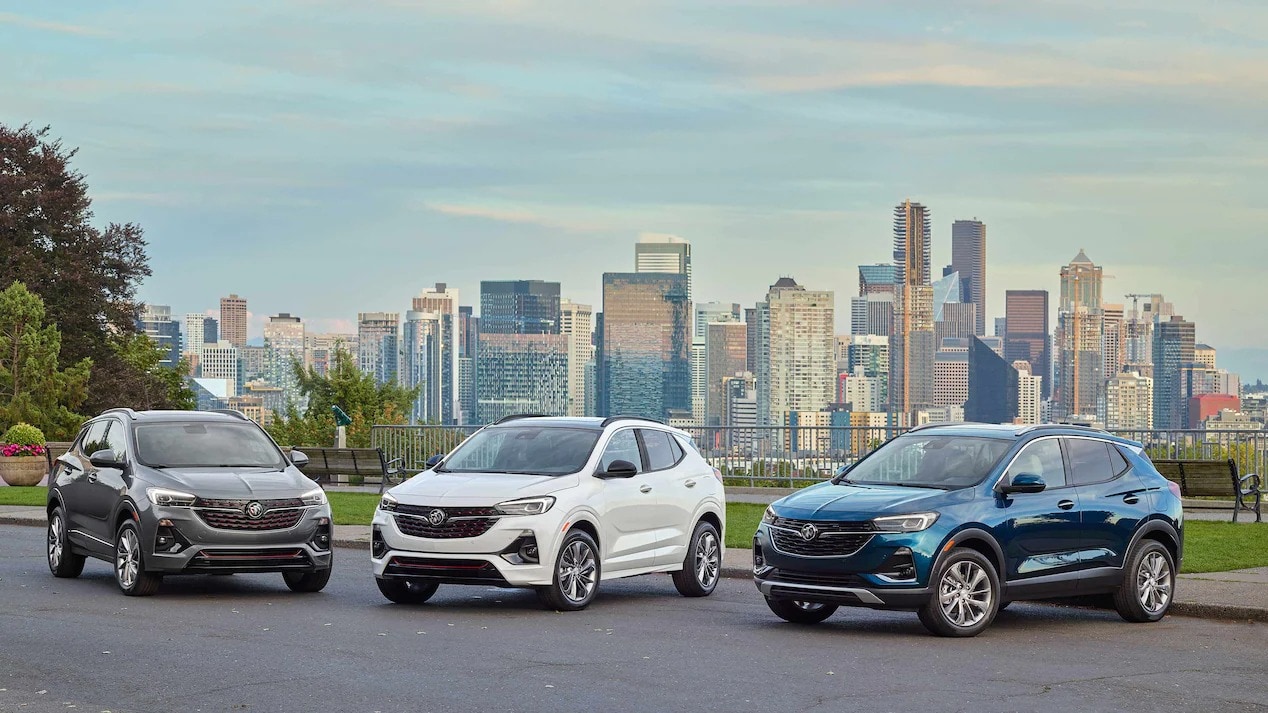 A grey 2023 Buick Encore GX on the left, a white one in the middle, and blue one of the right. All 2023 Buick Encore GXs are in front of a park in a city landscape