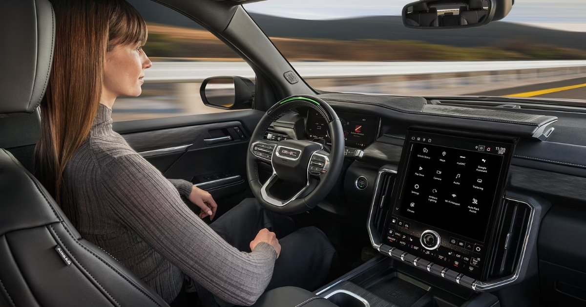 Woman seated in the driver’s seat of a 2026 GMC Acadia using hands-free Super Cruise driving technology, with a green light on the steering wheel and a large vertical touchscreen displaying vehicle apps and controls.