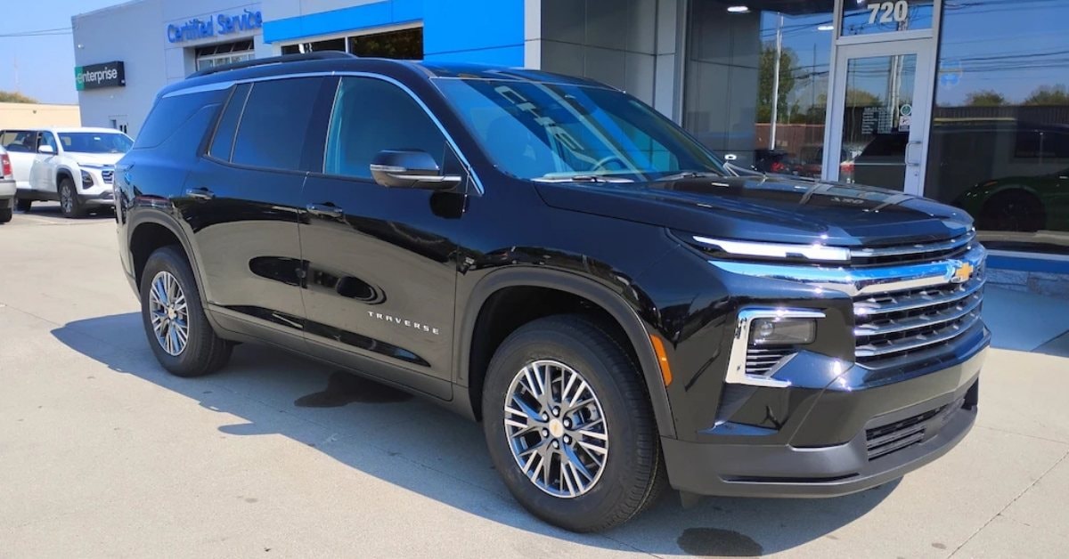  A shiny black Chevrolet Traverse SUV parked outdoors on a sunny day at a dealership or service center. The vehicle features a glossy black finish, chrome accents, and distinctive LED lights. In the background, a building with 