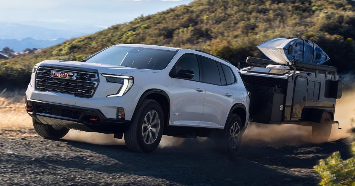 White 2026 GMC Acadia AT4 SUV driving on a dusty dirt road while towing a black off-road camping trailer with a kayak on top, set against a rugged sunlit hillside landscape.