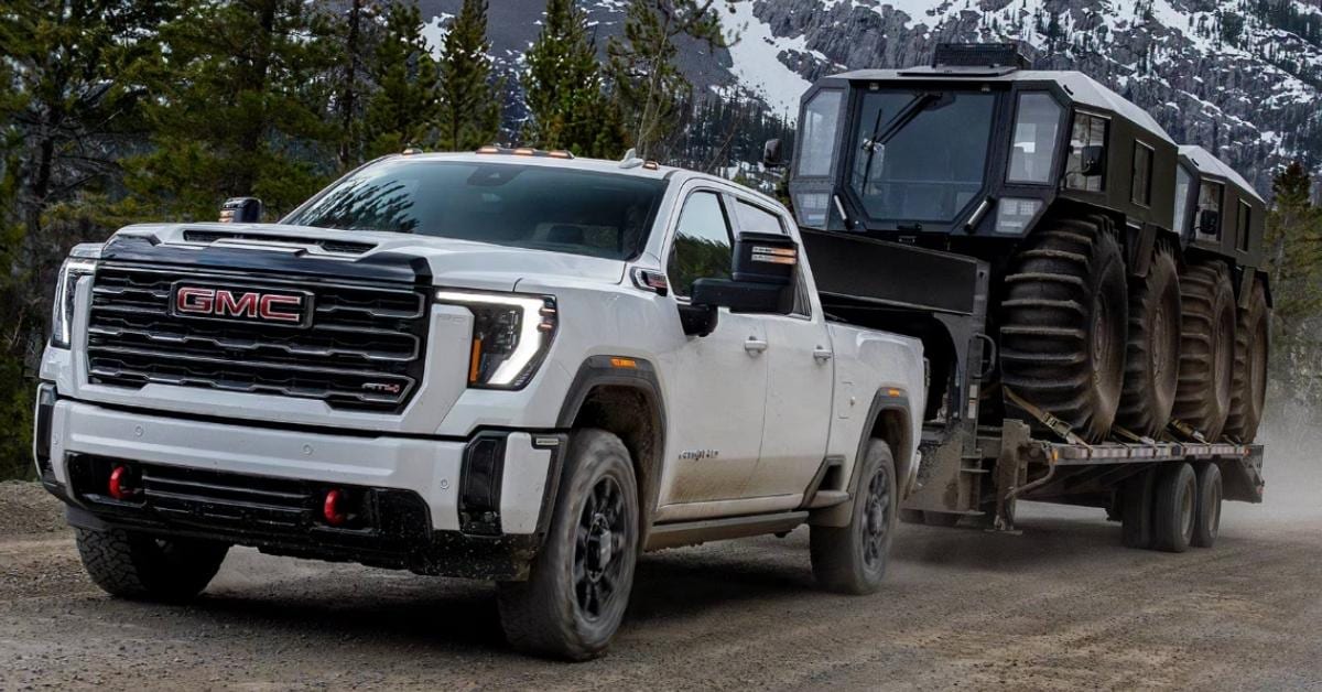  A white 2026 GMC Sierra 2500 HD AT4 pickup truck towing a heavy-duty flatbed trailer carrying a large black amphibious all-terrain vehicle. The truck features black trim, black wheels, and red recovery hooks, with dust on the tires and lower panels indicating off-road use. The scene is set on a dirt road surrounded by evergreen forests and snow-dusted mountains, showcasing the truck's towing power and rugged capability.