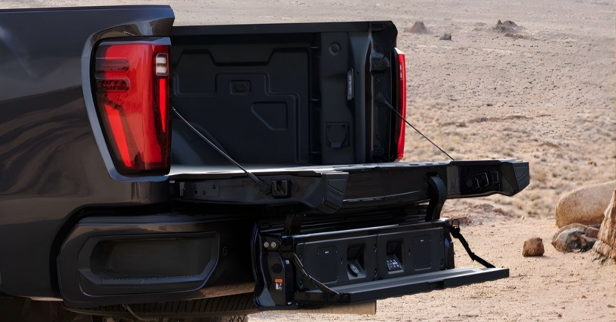  A close-up view of a 2026 GMC Sierra 2500 HD's multi-functional tailgate system, shown in a step configuration. The tailgate features a horizontal main section and a vertically lowered inner gate with a fold-out step. The truck bed is lined with a matte black finish and includes an LED bed light and a power outlet. The truck's dark metallic gray body contrasts with vibrant red LED tail lights and a rugged desert background with sandy terrain and sparse vegetation. The image highlights the tailgate's versatility and utility for work and outdoor activities.