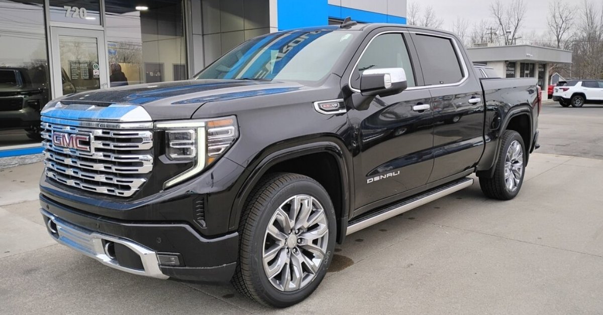 A black 2026 GMC Sierra 1500 Denali crew cab pickup truck with chrome accents, including a chrome grille, mirrors, and trim. The truck is parked on a concrete lot in front of a dealership, Don's Chevrolet GMC, with glass windows and blue accents. The 