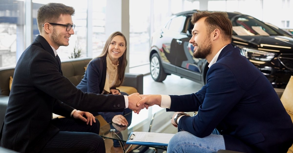 Two men shake hands across a glass table in a car dealership showroom while a woman smiles beside them, with an SUV in the background.