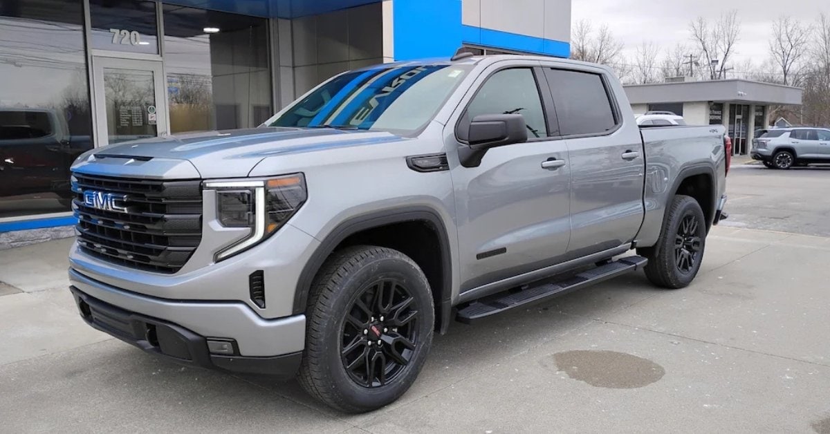 A silver 2026 GMC Sierra 1500 crew cab pickup truck with black accents, including a black grille, wheels, and trim. The truck is parked on a concrete lot in front of a dealership with glass windows and blue architectural accents. The overcast sky reflects on the truck's body.