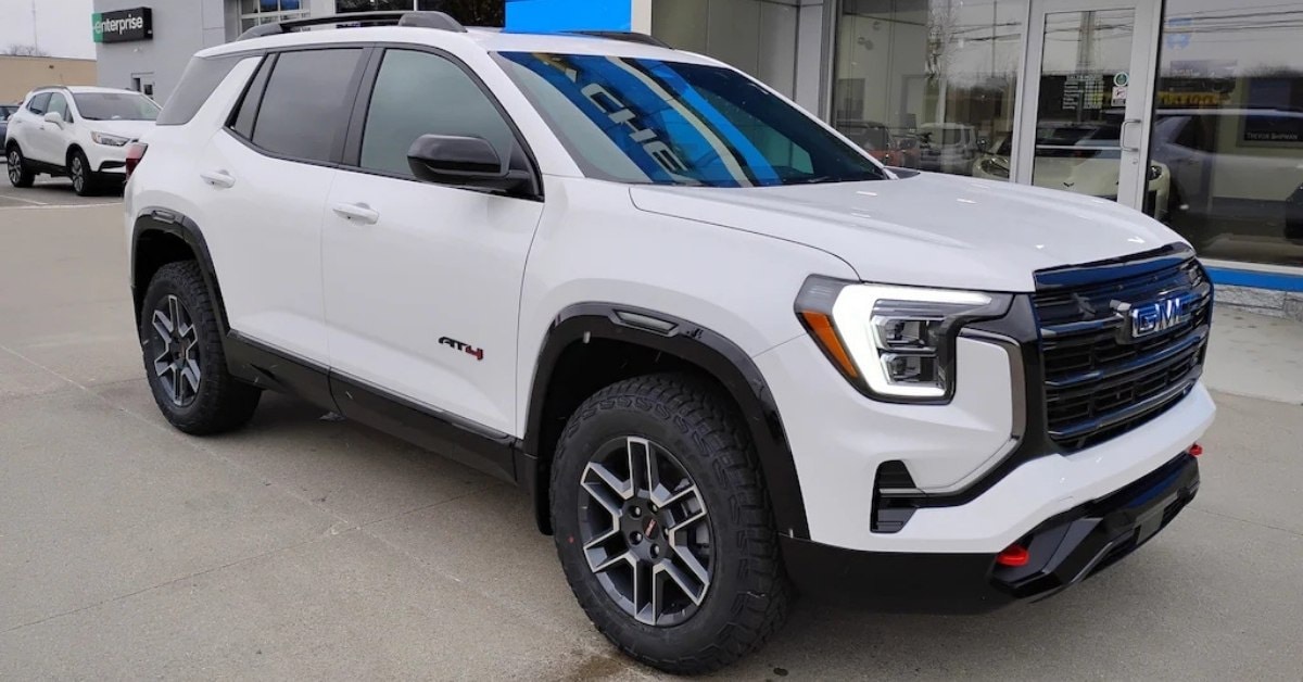  A white 2026 GMC Acadia AT4 SUV parked on a concrete surface in front of Don's Chevrolet GMC. The SUV features black accents, red tow hooks, and all-terrain tires. A green 'Enterprise' sign is visible on a nearby building, and reflections of 'Chevrolet' can be seen on the SUV's windshield and the dealership's glass windows.