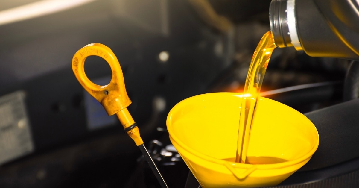  A close-up of fresh, golden engine oil being poured from a black plastic bottle into a bright yellow funnel positioned in a vehicle's engine bay. A yellow dipstick handle labeled 