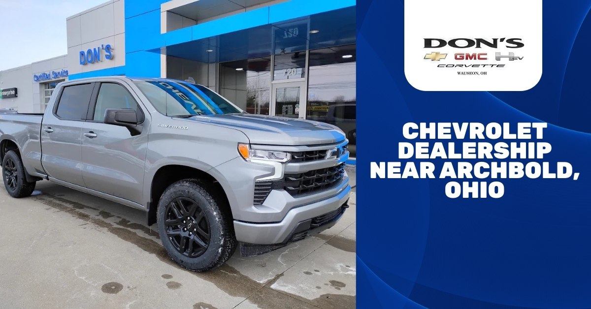  A silver Chevrolet Silverado truck is parked in front of a modern dealership building with 