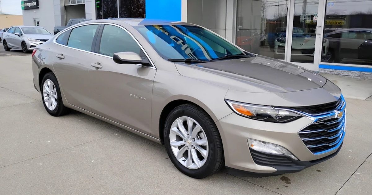  A metallic light gold Chevrolet Malibu sedan parked on a paved surface at a dealership or rental car lot. The car is angled slightly to the right, showcasing its front and left side. In the background, a building with an 