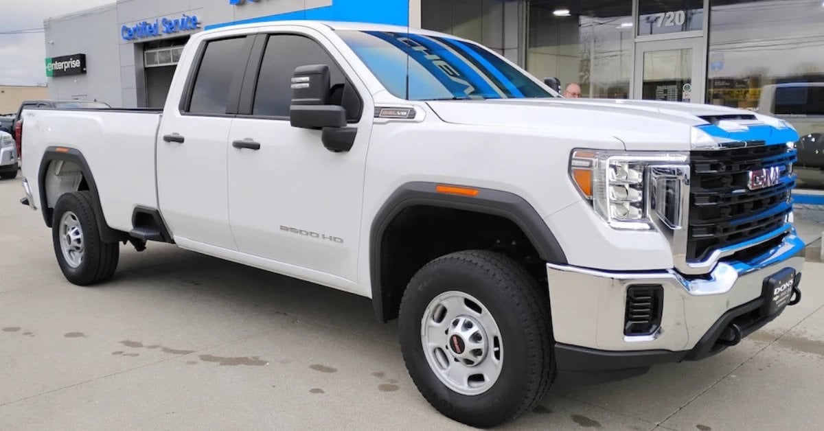 A white 2023 GMC Sierra 2500 HD pickup truck parked on a paved surface outside Don's Chevrolet GMC. The truck features a chrome grille, large side mirrors, black fender flares, and all-terrain tires. In the background, a building with 'Certified Service' and 'Enterprise' signage is visible, along with reflections of 'Chevrolet' in the truck's windshield. The setting suggests a professional automotive environment.