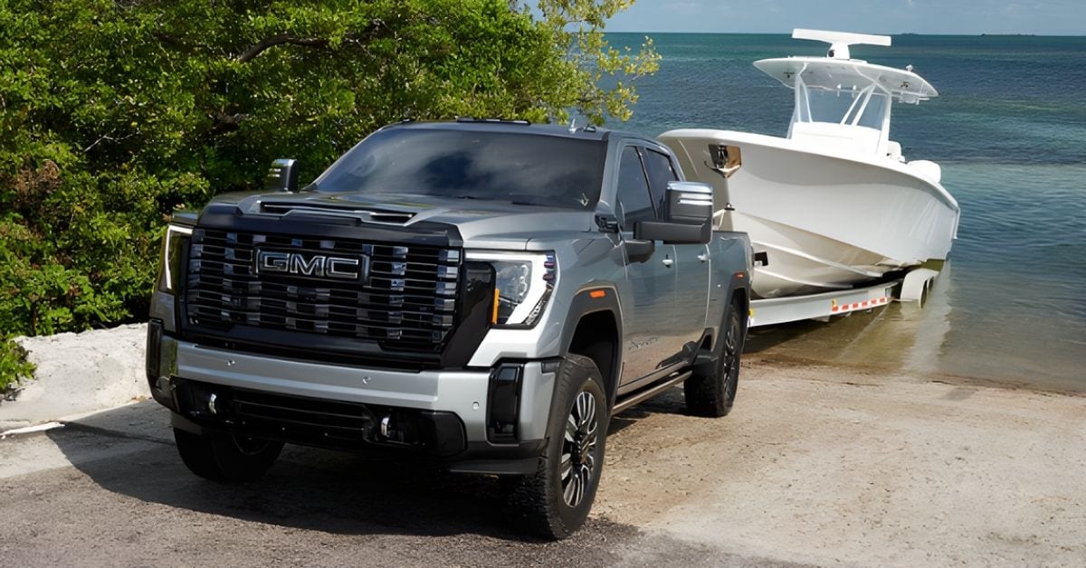  A silver 2026 GMC Sierra 2500 heavy-duty pickup truck towing a large white center-console powerboat down a concrete boat ramp into a calm, blue-green ocean. The truck features a chrome grille, chrome side mirrors, and black wheel-well trim, positioned diagonally in the foreground. The boat, equipped with a T-top and radar, is mounted on a trailer. The background includes lush green trees, a clear blue sky with light clouds, and the reflective ocean water, highlighting the truck's towing power and suitability for recreational activities.