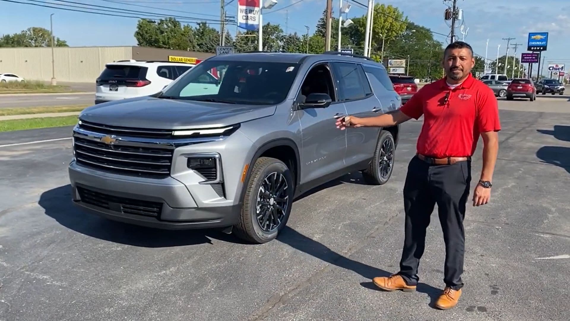  A man in a red polo shirt with a dealership logo stands next to a gray 2024 Chevrolet Traverse SUV in a parking lot, likely at Don's Chevrolet GMC. He is gesturing with one hand toward the vehicle, smiling at the camera. The SUV has a black grille, LED headlights, black wheels, and a roof rack. In the background, other vehicles and dealership signs are visible, including a 