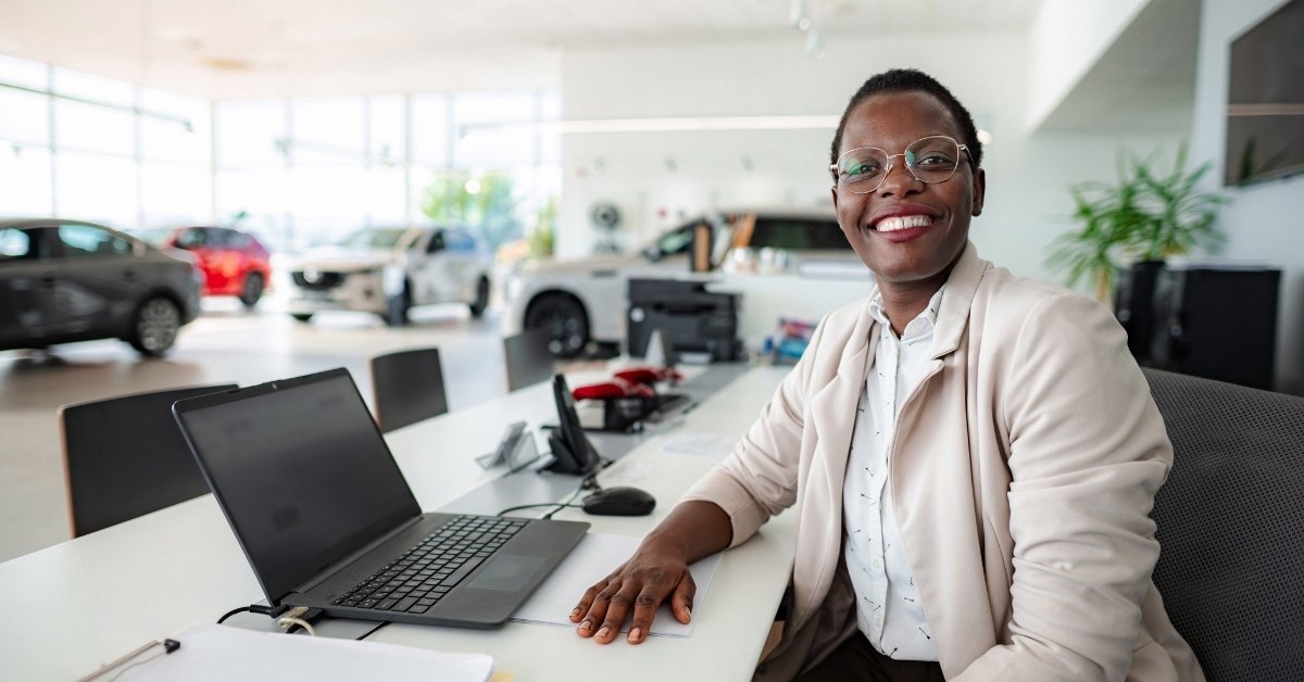 Smiling dealership sales representative seated at a desk with a laptop inside a bright car showroom, with several vehicles visible in the background.